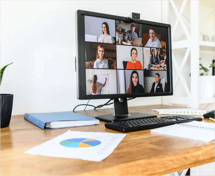 Desktop monitor showing a virtual team meeting with professionals participating via video call. The workspace features charts and office supplies, highlighting a remote work setting and digital collaboration environment